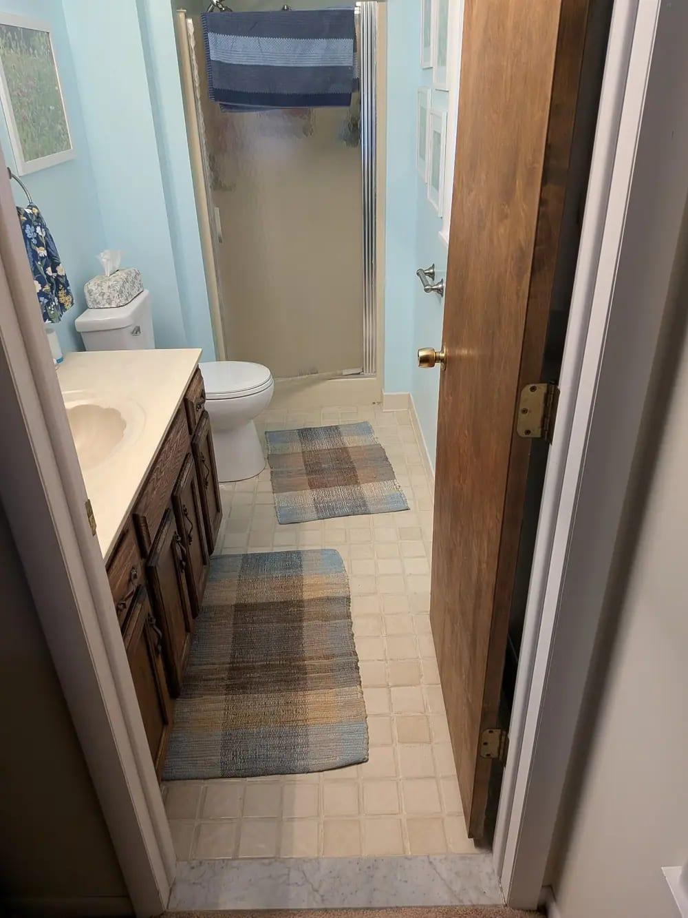 A small bathroom with a fiberglass shower stall, oak vanity and vinyl floor, before remodeling