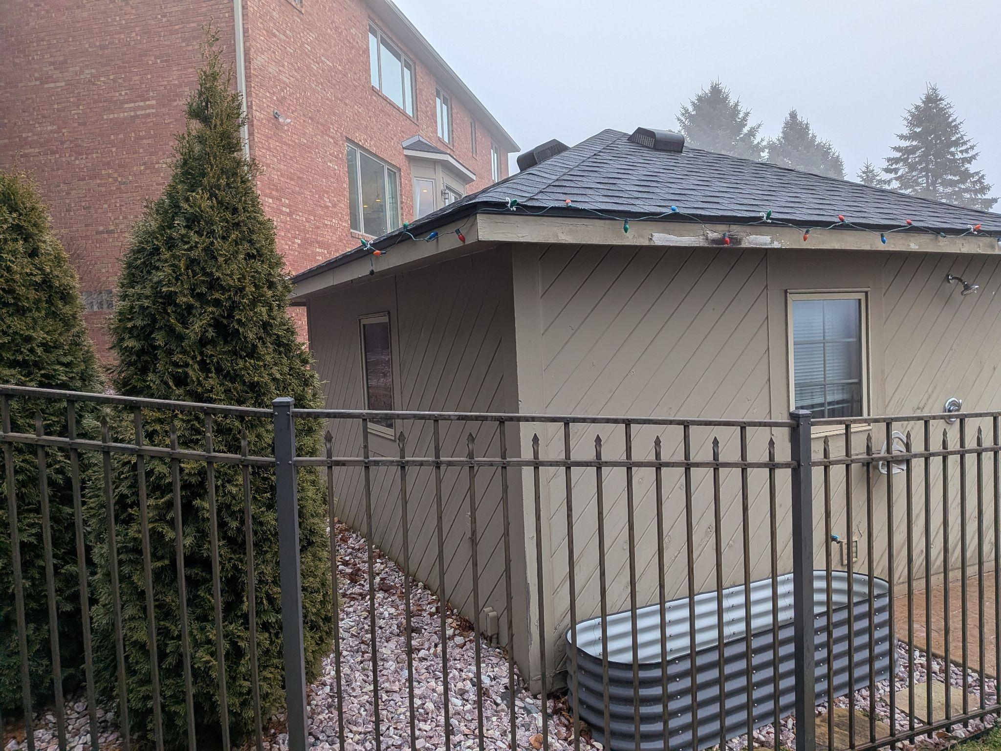 A small pool house with old tan diagonal siding and stripped roof trim, behind an iron fence