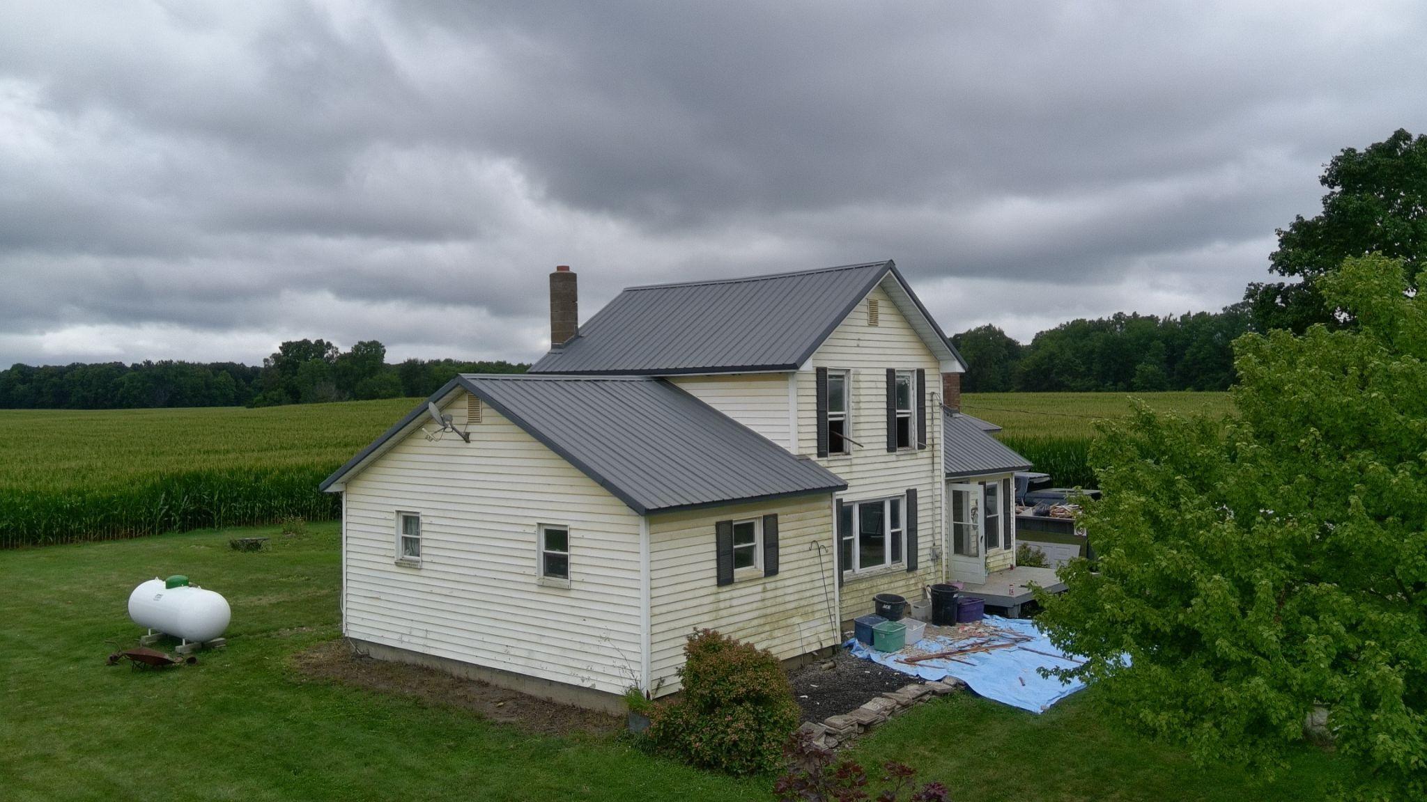 Aerial view of a two-story white farmhouse with a standing-seam metal roof and brick chimney, tarps on the ground