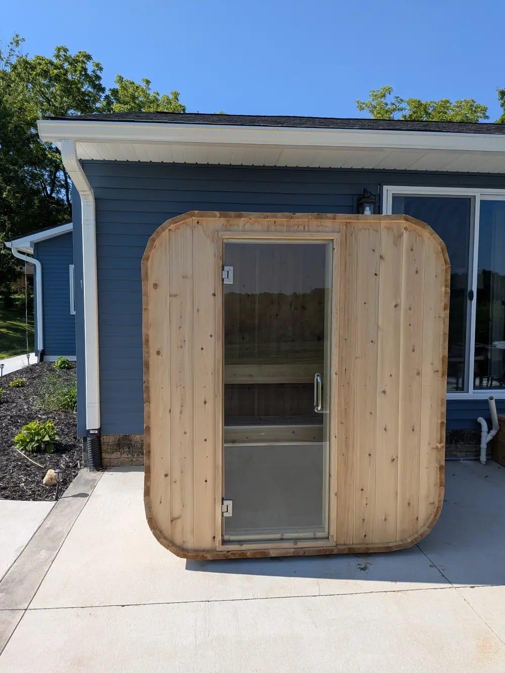 A cedar outdoor sauna with a glass door on a concrete patio beside a blue house