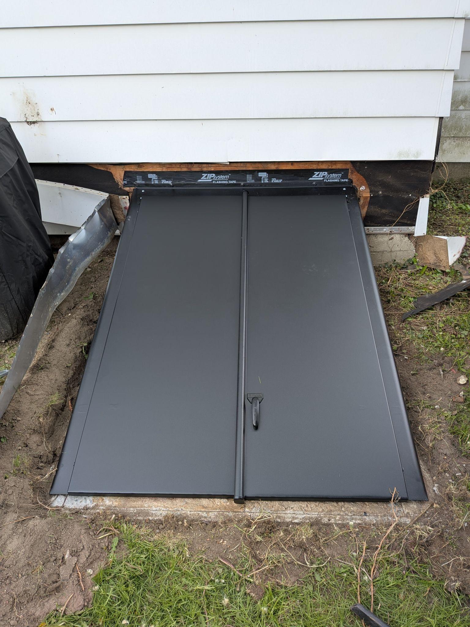 A steel basement egress door set into a window well beside a white house