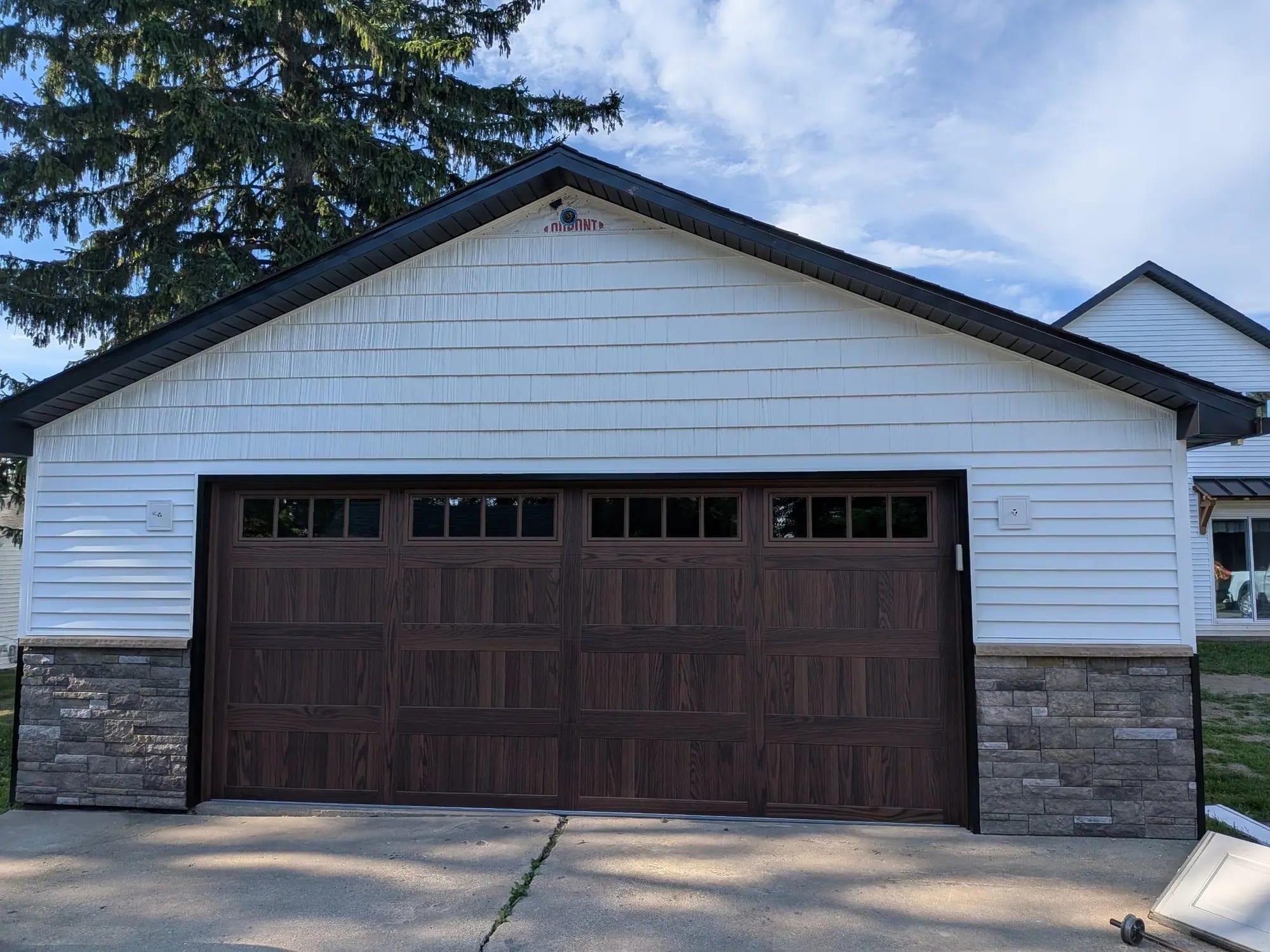 A two-car garage with white siding, stone veneer and a carriage-style wood-look door